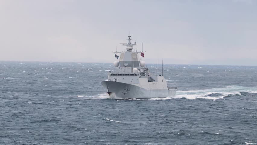 Modern naval warship cuts through rough ocean waves under cloudy sky during high speed maritime patrol mission.