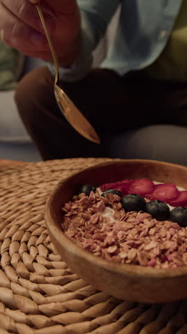 Vertical tilt up slowmo close up shot of bearded young Caucasian man eating yogurt and granola bowl with berries using spoon, enjoying delicious healthy breakfast on couch at home