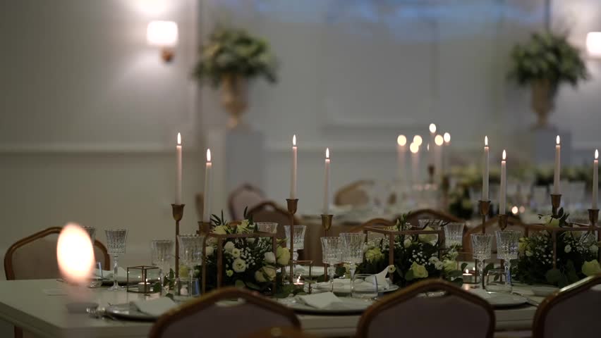 Long banquet dining table with gold candelabras, lit taper candles, white floral garland, crystal glassware and place settings in white ballroom