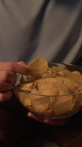 Vertical tilt up slowmo shot of bearded young Caucasian man enjoying bowl of delicious crispy potato chips while relaxed at home after work