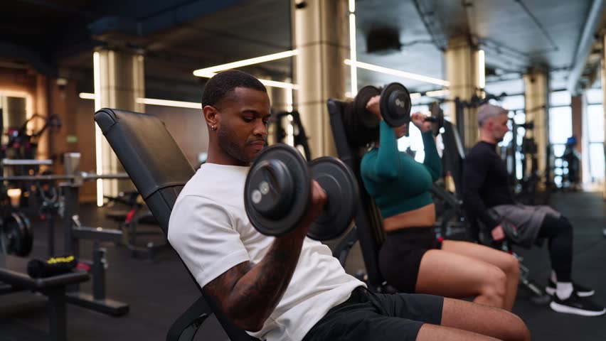 Young athletic man with tattoos lifting heavy dumbbells, focusing on his bicep curl workout. Diverse group of people training in a modern gym
