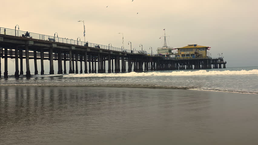 Santa Monica Pier stretches above calm ocean waves and wet sand, Moody coastal morning scene with reflections beneath historic wooden structure