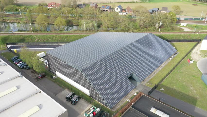 Aerial view of datacenter covered in solar panels, juxtaposed against green grass, a canal, and railway lines, Oostkamp, Flanders, Belgium.