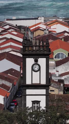 Aerial view of the dark stone clock tower amid the colorful roofs of Ribeira Grande with the ocean waves crashing in the background, Ribeira Grande, Portugal.