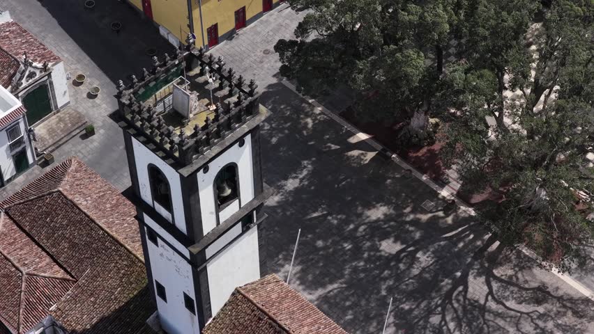 Aerial view of a tall church tower with black and white accents casting shadows amidst the old buildings and lush trees, Ribeira Grande, Portugal.