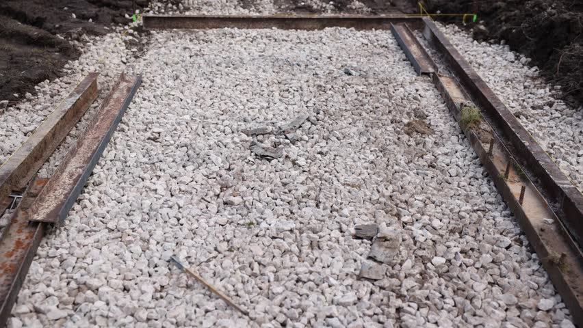 Close low angle view shows rusted rails, wooden stakes, and crushed limestone. Yellow string marks alignment, with soil edges showing fresh excavation work.