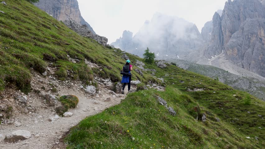 Cinematic shot of a hiker walking along a mountain trail surrounded by dramatic rocky peaks and misty fog. Atmospheric alpine landscape captured in the Dolomites, Italy. Ideal for travel, outdoor adventure, hiking, and exploration content in 4K resolution.