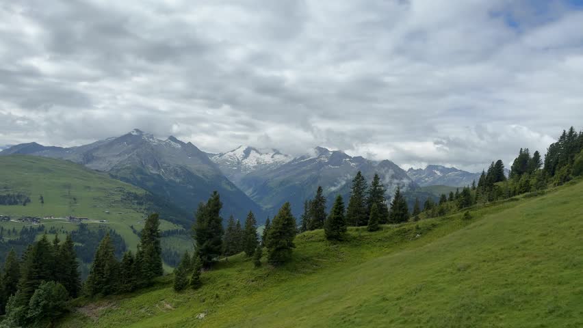 Panoramic summer scene of the Austrian Alps in the Salzachgeier range in a tracking shot, featuring lush green hills, deep valleys, and impressive snow-capped mountains under a cloudy sky. (Ultra HD)