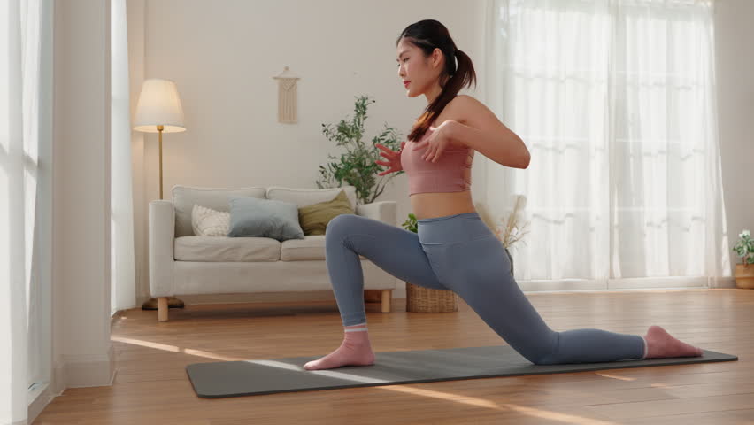 Asian woman in sportswear performing a lunging yoga pose with arms raised during her home workout session.