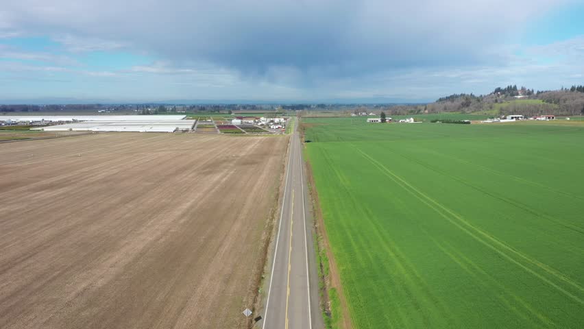 4K aerial drone footage of a rural road running through farmland in Oregon, USA under dramatic stormy clouds. Scenic countryside landscape shows cultivated fields on both sides with agricultural facility in the distance. Wide cinematic view highlights land use, rural environment, and changing weather conditions in the Pacific Northwest. Ideal b roll for travel, farming, transportation, and nature content.