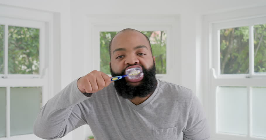 African American man bringing blue brush to mouth at home, brushing foamy teeth for oral care. Beard, grooming, toothpaste, bathroom, window, greenery, light