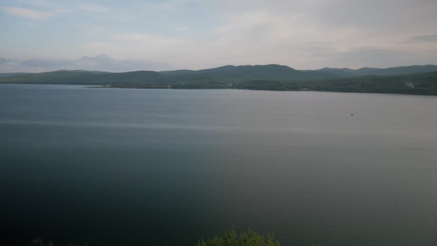 Static handheld top view overlooking a vast, calm mountain lake. Rolling green hills and a peaceful, cloudy sky stretch across the scenic background.