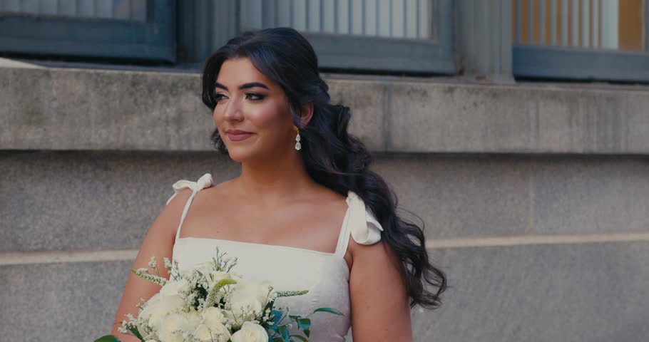 A close-up of a beautiful smiling bride in front of a building, as the groom gently touches her shoulder and kisses her.