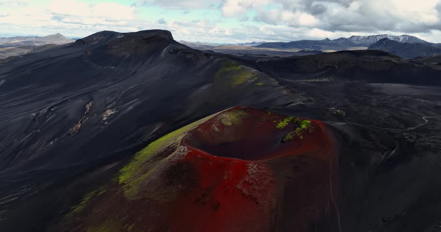 Aerial view of a vivid red volcanic crater surrounded by black ash mountains and bright moss patches, capturing stark color contrast and raw remote highland geology.