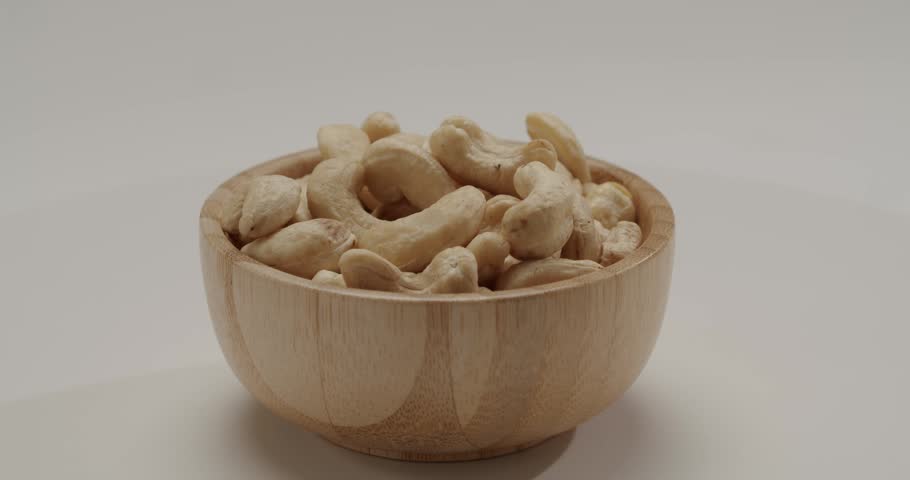 Rotating small wooden bowl filled with raw cashew nuts isolated on a white background, presenting a healthy and nutritious snack option for a balanced diet and vegan lifestyle.