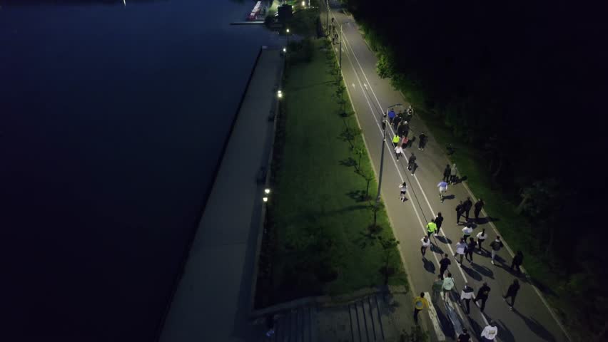Aerial view of a large crowd of people participating in a night running marathon along an illuminated lakeside alley, promoting an active and healthy lifestyle through sport and competition