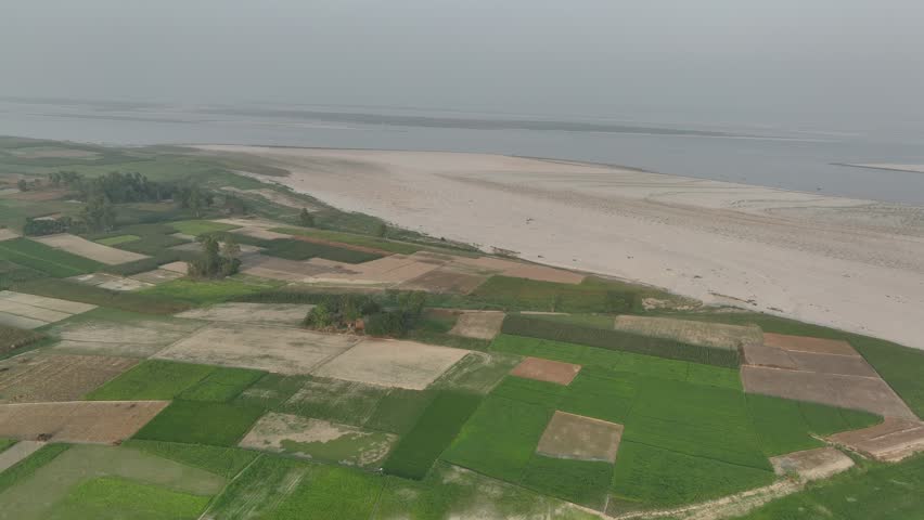 Stunning aerial footage flying over rural farmlands with vibrant green cultivated fields alongside a wide, sandy riverbank in a vast, hazy landscape under an overcast sky