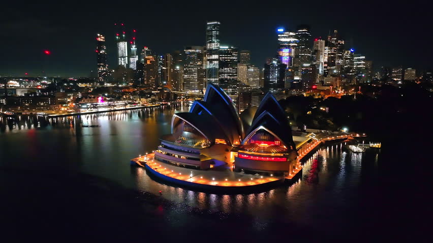 Australia, Sydney Opera House illuminated with vibrant lights at night, sparkling city skyline reflecting on calm harbor water, showcasing urban city life and travel background. Aerial flight panorama