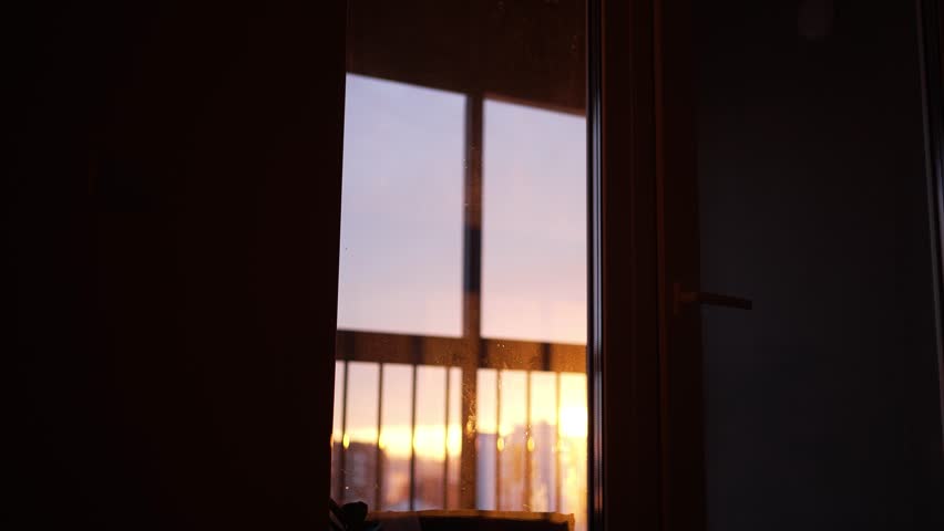 Golden sunlight from a setting sun moves across a glass door, revealing a balcony railing and casting a reflection with warm lens flares inside a dark, domestic interior