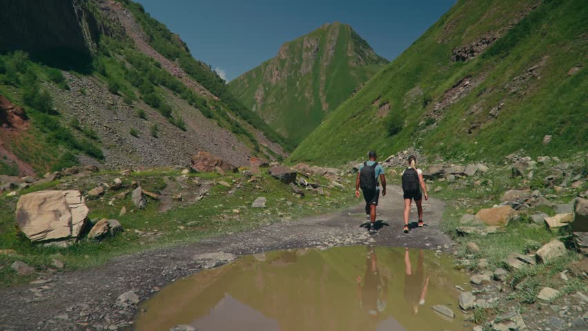 Two hikers walk along a gravel path with reflection in a puddle, surrounded by green mountains in Georgia, capturing peaceful nature and scenic outdoor adventure.