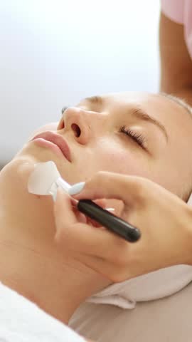 Beautician applies a gel peeling mask with a brush to a client’s face during a professional skincare treatment in a beauty clinic. Close-up cosmetology procedure for exfoliation, hydration skin