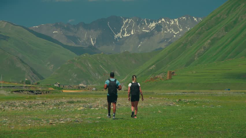 Two hikers walk in remote Truso Valley, Georgia, toward a snow capped mountain and fortress ruins, surrounded by green mountains and blue sky, capturing adventure and history.