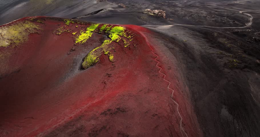 Aerial view of a red volcanic crater slope in Iceland with neon green moss and a zigzag hiking path, capturing surreal geology, raw highland color, and remote wilderness.