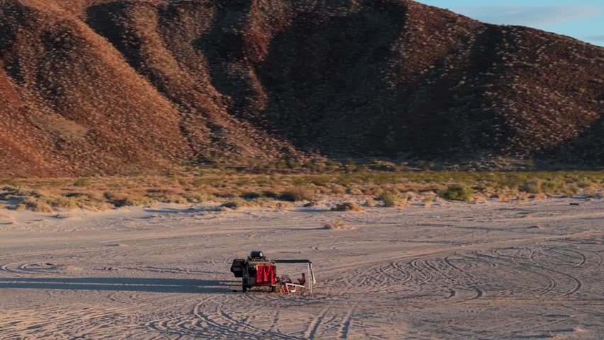 View of an SUV and teardrop camper parked on the beach in Baja California Mexico while the sun rises or sets over the desert landscape.