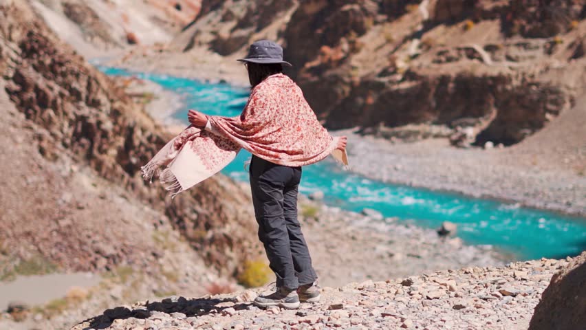 Teenager girl wearing cap standing near mountain river while enjoying beautiful views of blue color water of Tsarap river in Zanskar, Ladakh, India. Tourist exploring hidden location in remote Ladakh