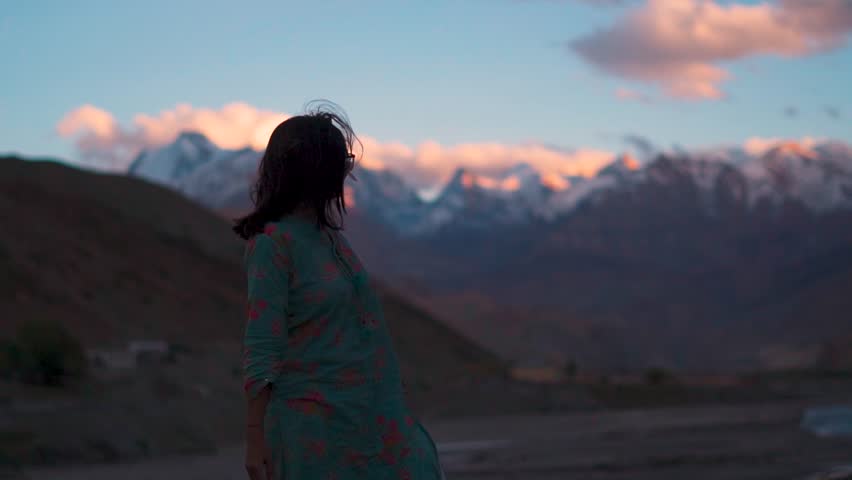 Silhouette of teenager girl raise hands against snowy mountains during sunset at Spiti Valley, Himachal Pradesh, India. Trekker breathing fresh air on the top of a mountain. Travel and enjoying life.