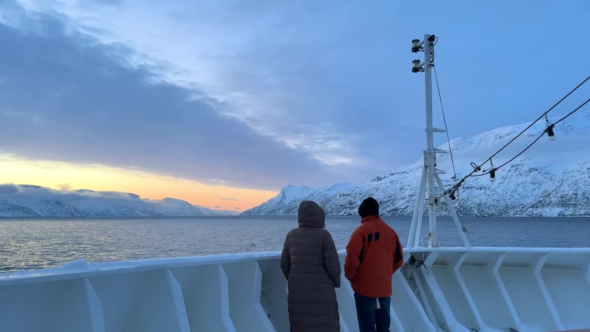 People stand on the deck of a cruise ship in Norway. They watch the mountains and ocean as the sun sets. Snow is present on the ground and the air feels cold.