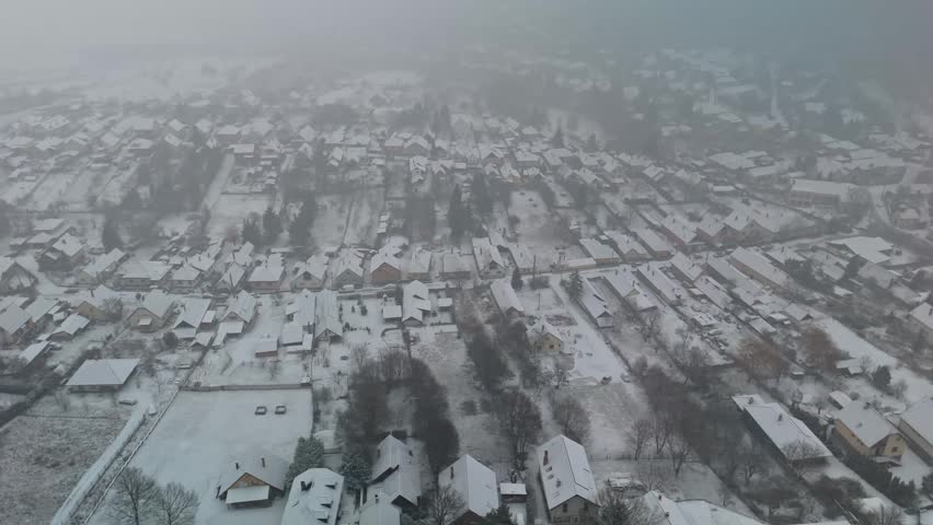 Pilisszentlászló village in the Pilis Mountains features snow-covered rooftops, frosted trees, and winding roads amid the Hungarian countryside, captured from a sideways drone perspective in winter.