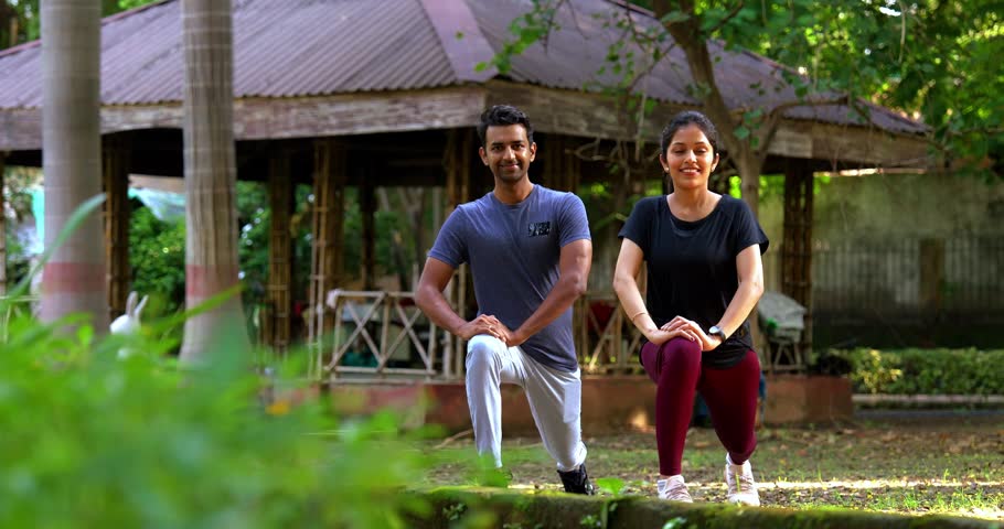 Indian beautiful young couple stretching legs while standing post fitness in park during sunny morning outdoor session, focusing on flexibility, health, wellness, active lifestyle motivation together