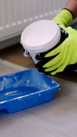 House Painter in gloves pours white paint from Can into a blue tray At Living Room Renovation Painting