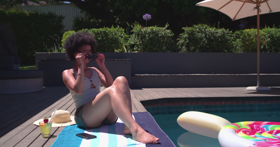 Woman is donning sunglasses under sun at pool edge, sipping drink as camera is panning. Relaxation, leisure, outdoor, backyard, deck, towel, shades