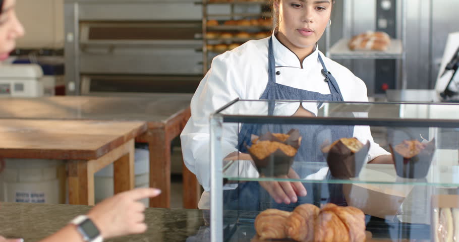 Customer pointing at case, baker in apron grabbing muffin with tongs, packing for purchase at shop. Bakery, pastry, display, counter, chef, packaging, takeout