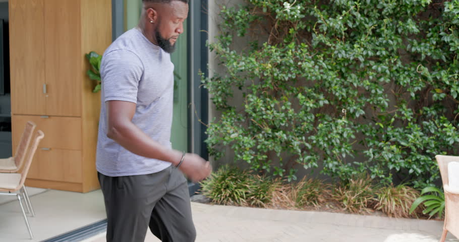 African American man exiting sliding door to patio with towel shielding eyes drinking green bottle. Outdoor, hydration, stretch, fitness, sunlight, smartwatch, plants