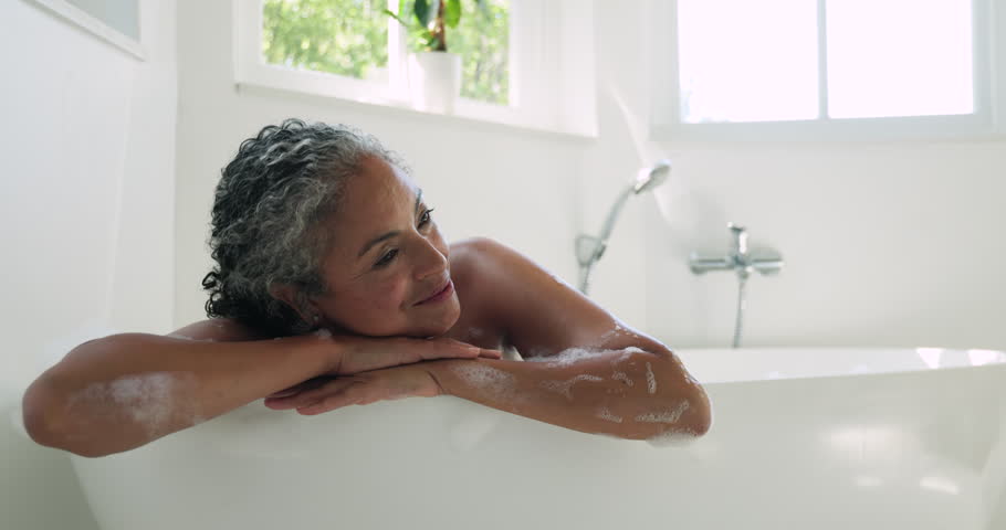Senior woman leaning on tub rim, suds, camera zooming, smiling and laughing at lens in bathroom. Bathtub, minimalist, bright, natural, selfcare, serenity, wellness