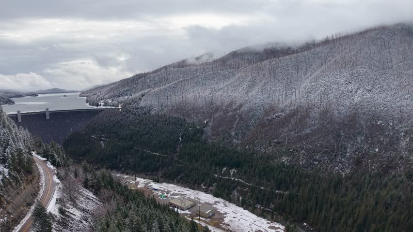 Aerial view of a snow-covered dam and winding road nestled between evergreen mountains near Flathead Lake in Montana under a cloudy winter sky.