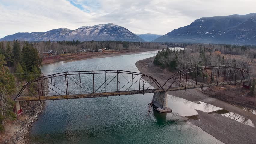 Aerial view of an old metal bridge spanning a turquoise river with mountains and forested banks near Glacier National Park in Montana