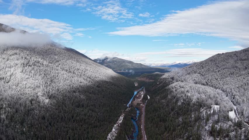 Aerial drone footage of snow covered evergreen mountains and forested valley near Whitefish Montana with a winding blue river and expansive winter landscape under a bright sky