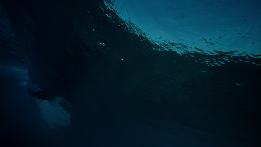 Underwater ocean beneath wave with dark blue water and surface ripples, rearview follows shimmering surfer silhouette riding wave, behind pov