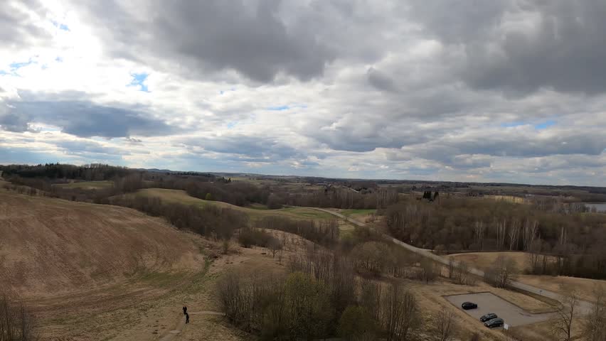 Wide countryside landscape with lake, trees, and a house under a dramatic cloudy sky.