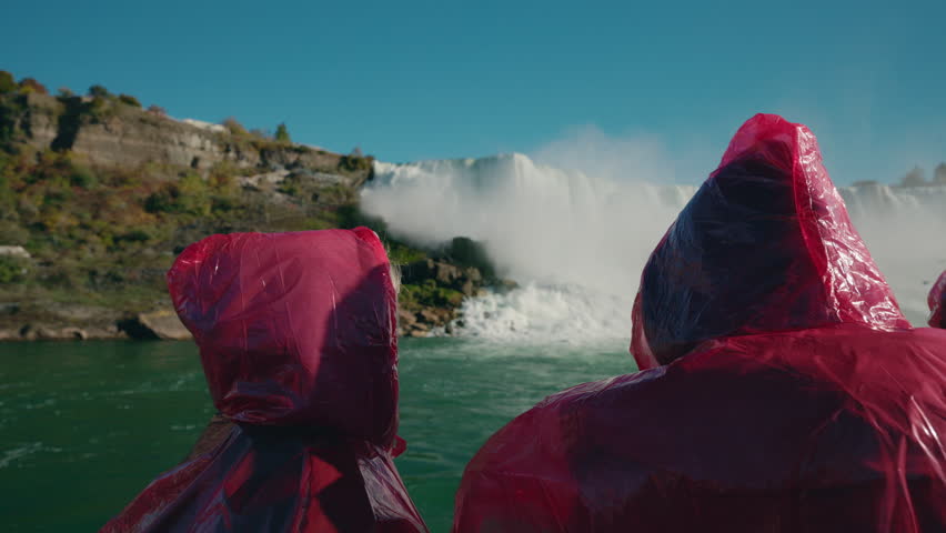 Tourists wearing red rain ponchos watching Niagara Falls waterfall from boat close to the cascade.