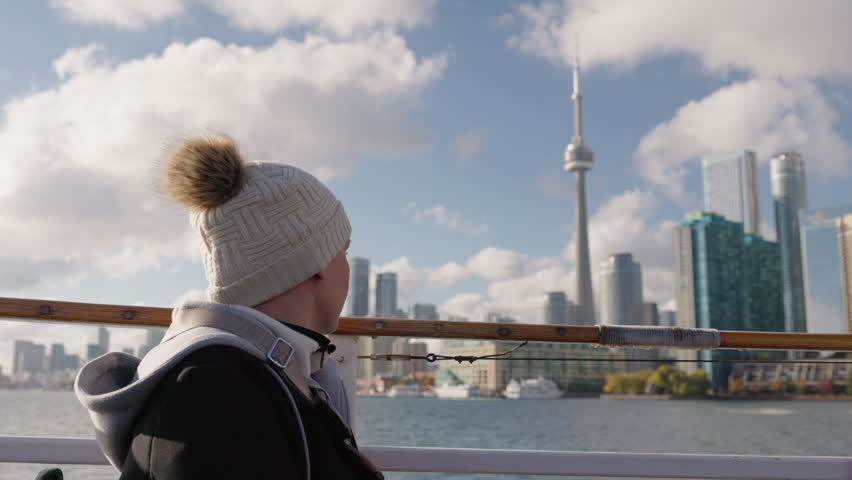 Teenage girl passenger looking at Toronto skyline from boat on Lake Ontario, side view with winter hat and calm travel atmosphere on waterfront.