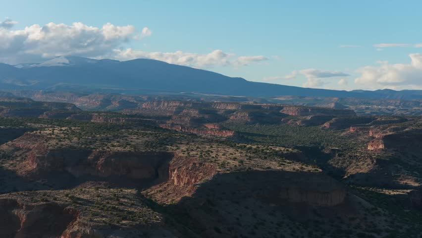 A cinematic drone shot captures expansive southwest mesa plateaus and rugged mountains. Dramatic clouds on the horizon fill the blue sky over this vast and universal high desert wilderness.