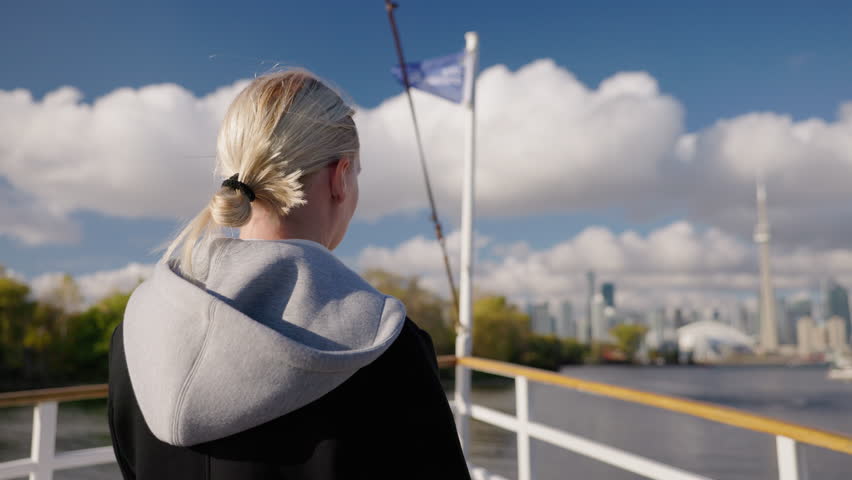 Young woman tourist on boat deck looking at Toronto skyline across Lake Ontario, back view with blonde hair and calm travel atmosphere on waterfront.