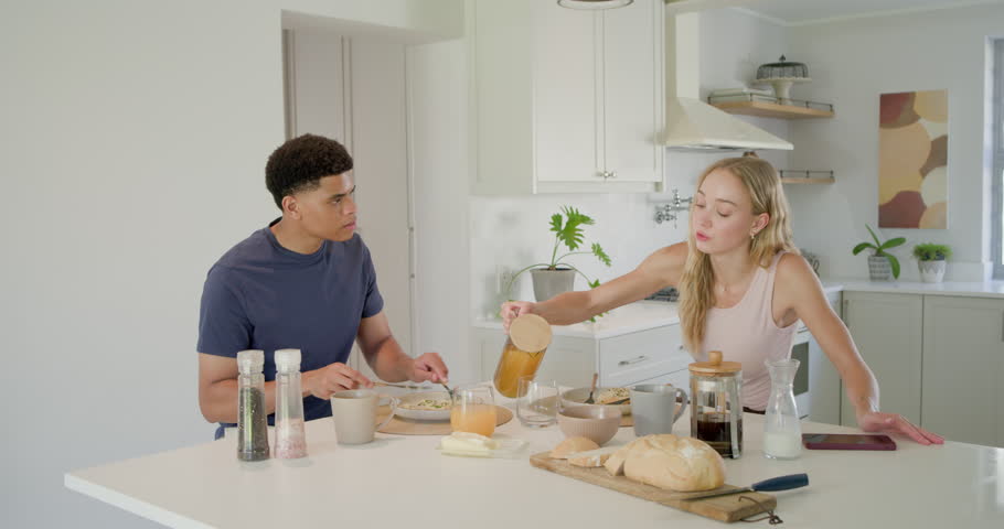 Couple starting breakfast at kitchen island as woman pouring orange juice, man cutting bread. Duo, home, morning, bright, modern, countertop, carafe