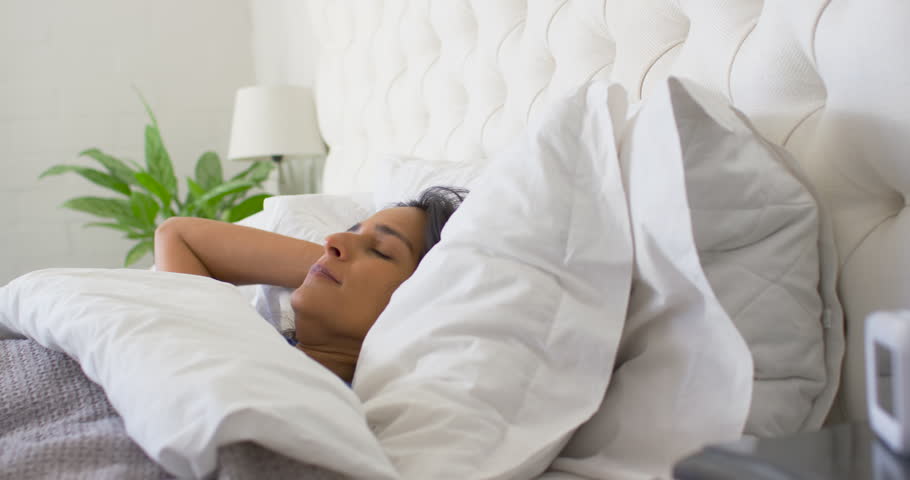 Indian adult woman wearing blue sleepwear waking in bed to white alarm clock ringing and silencing. Bedroom, morning, sunlight, duvet, pillows, plant, lamp