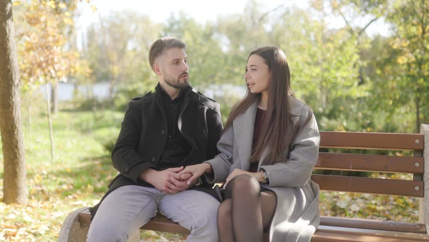 A lovely young romantic couple sits comfortably on a wooden park bench during a beautiful autumn day. They lovingly hold hands while chatting and resting together.